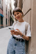 © PhotoBook - Close up outside photo of smiling cute lady using smartphone while posing near the wall outdoor