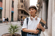 © PhotoBook - Cute pretty girl with light short hair is holding notes and backpack posing to camera outside