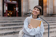 © PhotoBook - Cute charming young woman with wonderful happy smile is holding notes and looking to camera while posing in front of university