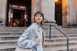 © PhotoBook - Stylish young woman with light hair wearing grey shirt and holding backpack is looking aside in front go university