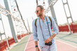 Young attractive man with photocamera exploring a new city with a backpack behind by walking outdoors. Tourist walking on the bridge sightseeing having fun