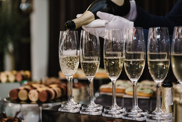 The waiter pouring white sparkling wine. Catering service concept. Barmen pours champagne into flute glasses. Champaign is being poured into glasses. Bottle in a closeup view. Rows of full glasses.