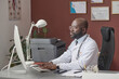 © AnnaStills - African american doctor in white lab coat sitting at his desk in office and working on computer