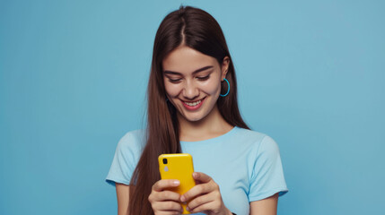 Canvas Print - smiling young woman looking down at her yellow smartphone with pleasure, wearing a blue top and matching blue hoop earrings against a soft blue background