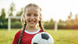 © Studio Nova - happy young girl with braided hair, holding a soccer ball, wearing a red sports jersey, with a soccer goal in the background, likely on a playing field