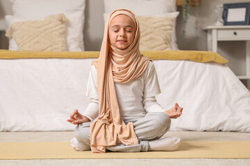 Wall Mural - Little Muslim girl in hijab meditating on yoga mat at home