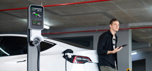 Young man travel with EV electric car to shopping center parking lot charging in downtown city showing urban sustainability lifestyle by green clean rechargeable energy of electric vehicle innards