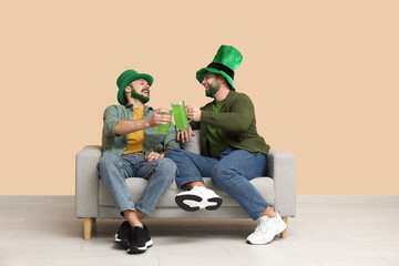 Wall Mural - Young men in leprechaun hats with green beards holding glasses of beer and sitting on sofa near beige wall. St. Patrick's Day celebration