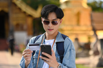 A handsome Asian tourist using a credit card and smartphone for cashless payment online for travel, Thai Temple on background