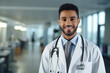 © byerenyerli - A young male doctor wearing a white coat and stethoscope has a reassuring smile. A hospital and clinic environment with medical equipment visible in the background that reflects trust and professional