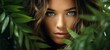 Beautiful young woman's face with natural makeup and green eye behind green leaves while looking at the camera