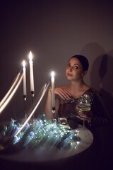 Portrait of a girl in a gold festive sequined dress sitting at a round table at home on Christmas night. On the table there are glasses with wine and branches of fir tree with garland.