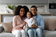 © Home-stock - Cheerful black family of three people hugging sitting on couch at home, posing for photo and smiling at camera