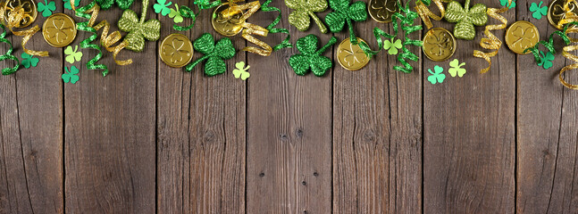 Poster - St Patricks Day top border of green shamrocks, gold coins and ribbon. Above view over a rustic dark wood banner background with copy space.