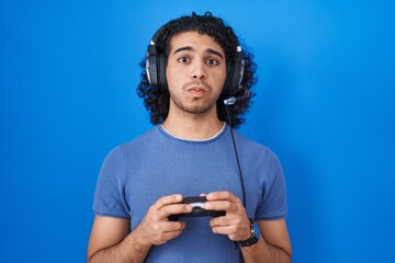 Canvas Print - Hispanic man with curly hair playing video game holding controller puffing cheeks with funny face. mouth inflated with air, catching air.