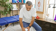 Mature hispanic man holding knee with pained expression in a rehabilitation clinic.