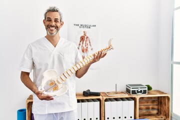 Poster - Middle age grey-haired man physiotherapist smiling confident holding anatomical model of spinal column at rehab clinic