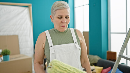 Poster - Middle age grey-haired woman holding paint roller with serious face at new home