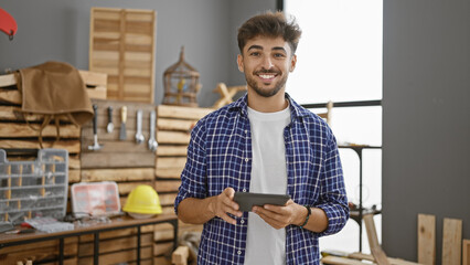 Wall Mural - Smiling young arab man takes on carpentry business, embracing technology with touchpad in his bustling indoor workshop