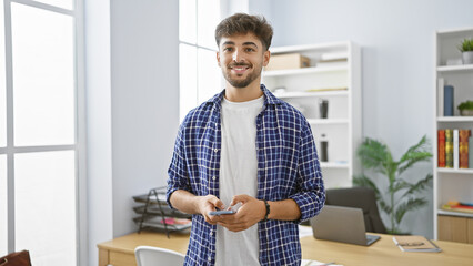 Canvas Print - Confident young arab man, beaming with a happy expression, engrossed in work, effortlessly manages business tasks on his smartphone, standing in an elegant office indoors.