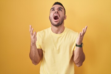 Canvas Print - Young hispanic man standing over yellow background crazy and mad shouting and yelling with aggressive expression and arms raised. frustration concept.