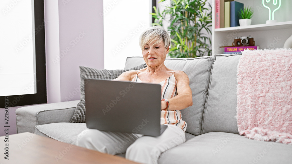 Mature woman with grey hair using a laptop on a sofa in a cozy living ...