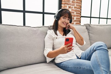 Wall Mural - Young beautiful hispanic woman listening to music sitting on sofa at home