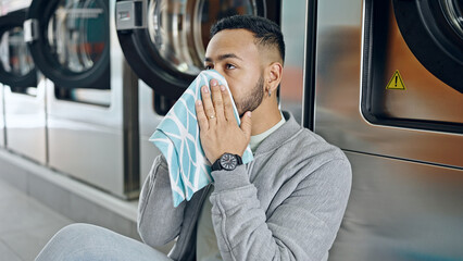 Wall Mural - Young hispanic man smelling cloth sitting on the floor at laundry facility