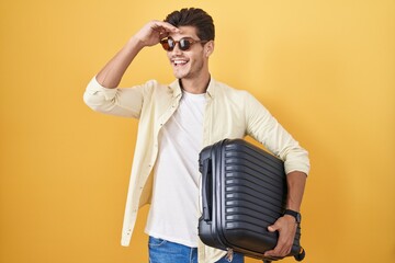 Wall Mural - Young hispanic man holding suitcase going on summer vacation very happy and smiling looking far away with hand over head. searching concept.