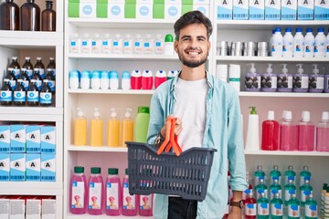 Sticker - Young hispanic man with tattoos shopping at pharmacy drugstore holding basket looking positive and happy standing and smiling with a confident smile showing teeth