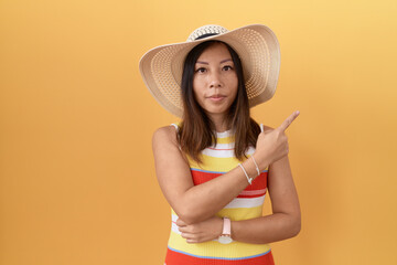 Wall Mural - Middle age chinese woman wearing summer hat over yellow background pointing with hand finger to the side showing advertisement, serious and calm face