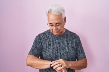 Wall Mural - Middle age man with grey hair standing over pink background checking the time on wrist watch, relaxed and confident