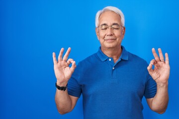 Canvas Print - Middle age man with grey hair standing over blue background relax and smiling with eyes closed doing meditation gesture with fingers. yoga concept.