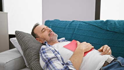 Poster - Sleepy middle age man with grey hair, engrossed in his book, succumbs to a relaxing snooze, comfortably lying on his cozy sofa at home.