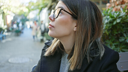 Wall Mural - A contemplative young adult hispanic woman wearing glasses sits at an outdoor cafe table.