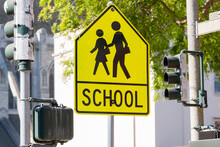 Yellow Warning Road Sign For A School Zone At A Road Intersection With A Street Traffic Light In The Background. Yellow School Zone Street Sign, Close-up. School Road Sign With Children, Foreground