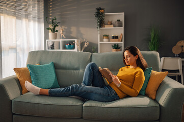 Wall Mural - Woman relaxing and reading a book at home while lying on a sofa