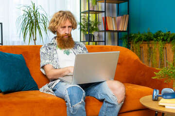Caucasian redhead man freelancer at home living room apartment and sits down on couch opens laptop. Young bearded guy works on notebook, sends messages, makes online purchases, watching movies