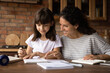© fizkes - Mother assist in learning. Caring hispanic mom help preteen daughter in doing homework check written task correct mistake. Young female teacher explain grammar rule to little girl pupil at home lesson