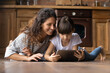 © fizkes - Friendly millennial hispanic mother and preteen daughter relax on wooden warm floor at kitchen play videogame on tablet device. Caring mexican nanny watch education channel with small girl on pad pc