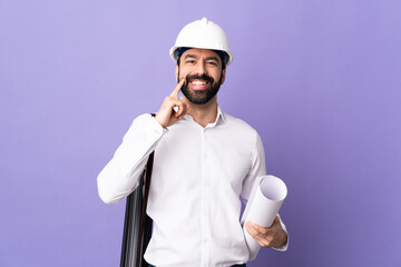 Wall Mural - Young architect man with helmet and holding blueprints over isolated purple background smiling with a happy and pleasant expression