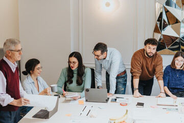 Wall Mural - A multigenerational group of men and women gather around a table, collaborating in a business meeting focused on strategy and innovation. Laptops, notes, and casual work attire suggest a modern