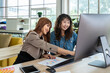 Two smiling young Asian female woman businessperson graphic designer working together, meeting and brainstorming, using computer in office, pointing at document.