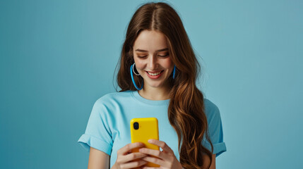 Poster - smiling young woman looking down at her yellow smartphone with pleasure, wearing a blue top and matching blue hoop earrings against a soft blue background