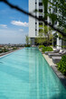 A serene rooftop swimming pool extends towards the horizon, bordered by modern lounge chairs and vibrant greenery, against a backdrop of cityscape and clear blue skies..