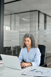 © insta_photos - Busy mature middle aged professional business woman, smiling older manager executive leader with gray curly hair looking at laptop computer in office working typing on pc sitting at desk. Vertical.