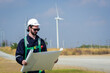 © pornchai - Handsome caucasian engineer, technician man wearing safety uniform workwear standing, holding and look at drawing paper or blueprint in front of windmill or turbines field with blue sky background.