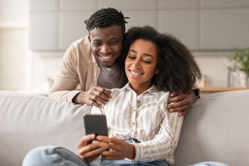 Wall Mural - Cheerful black couple sharing social media on cellphone at home