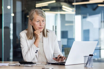 Focused mature female accountant working on laptop while sitting at workplace. Gray-haired employee woman in fashionable suit using portable computer for writing reports and checking email at office.