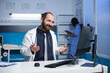 Bearded Caucasian man wearing a lab coat in an office engaged in a video call on a desktop PC, discussing medical matters. Other healthcare professionals work in the background.